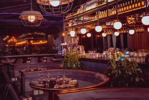 Interior de un restaurante y bar de ambiente sofisticado, con barra iluminada, botellas y mesas elegantemente dispuestas.
