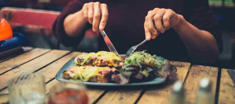 Persona disfruta un plato de comida en un restaurante, cortando una preparación acompañada de ensalada y otros alimentos.