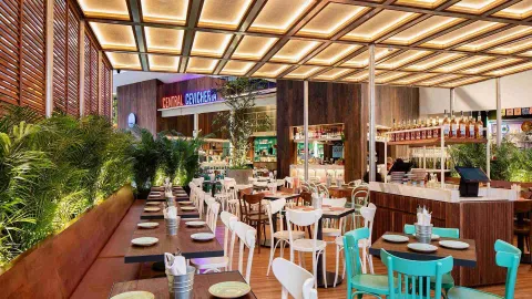 Amplio salón del restaurante con mesas de madera, sillas blancas y turquesas, plantas verdes y techo iluminado.