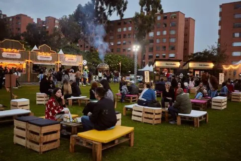 Evento gastronómico al aire libre, con asistentes disfrutando de comida, bebidas y música en un parque rodeado de edificios residenciales.