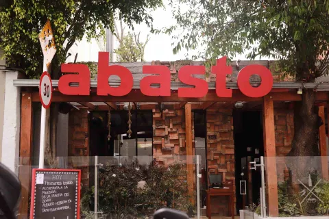 Fachada de Abasto con letrero rojo, estructura de ladrillo y madera, vegetación exterior y un tablero que anuncia productos locales.