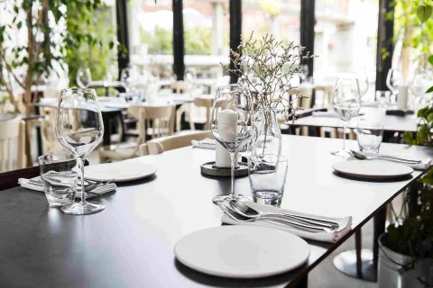 Mesa de restaurante elegantemente dispuesta con vajilla blanca, copas de vino, cubiertos y un arreglo floral, en un ambiente luminoso y rodeado de vegetación.