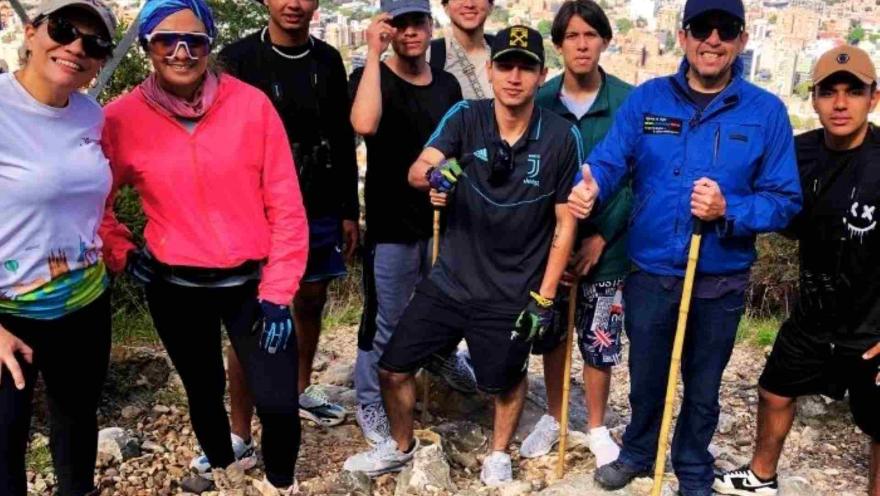  Grupo de personas jóvenes junto con un guía, delante de un mirador donde se observa la ciudad de Bogotá. Todos con ropa deportiva, algunos hombres llevan gorra.