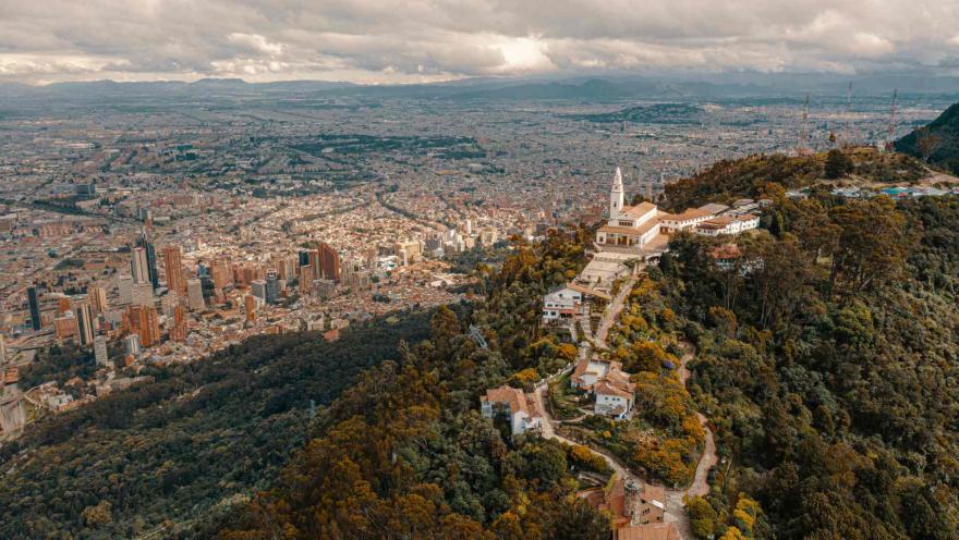 Monserrate Hill, a wide aerial shot taken by drone showing the entire complex surrounded by trees on top of the mountain. In the distance, the vast city and its buildings can be seen all the way to the chain of western mountains. The sky is overcast with clouds.