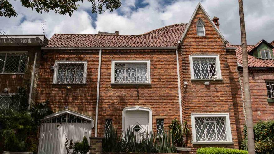 Chapinero primer plano fachada casa estilo inglés en ladrillo, techos en teja de barro y ventanas con marcos y rejas metálicas. La casa cuenta co antenjardín con plantas, asío como la puerta al garaje.