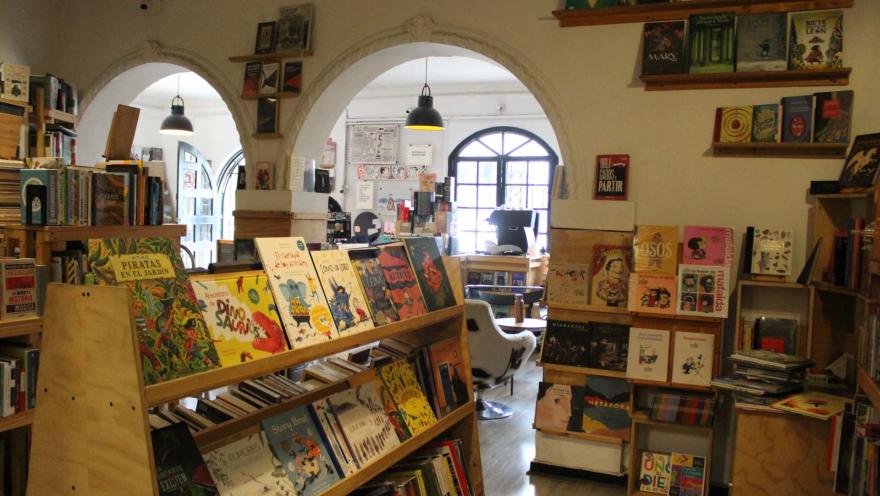  Garabato Libros salón con estanterías en piso y paredes abarrotadas con libros. Al fondo se observa otro salón con un ventanal con figuras geométricas y una puerta que conduce a la calle.