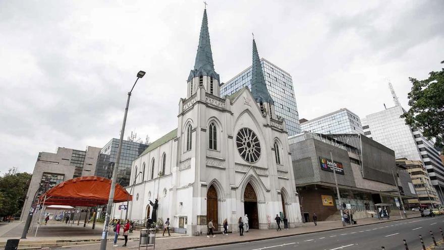 Iglesia La Porciúncula vista fachada gótica y blanca sobre la calle 72 delante de edificios modernos y una carpa para eventos. Varias personas caminan alrededor. Destacan las dos agujas del techo color gris en un ciuelo blanco.