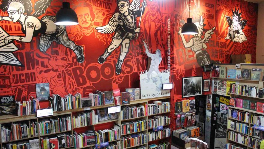 Librería La Valija de Fuego anaqueles repletos de libros bajo una pared roja con imagenes dibujadas en negro y blanco de personas con alas y libros en las manos.  