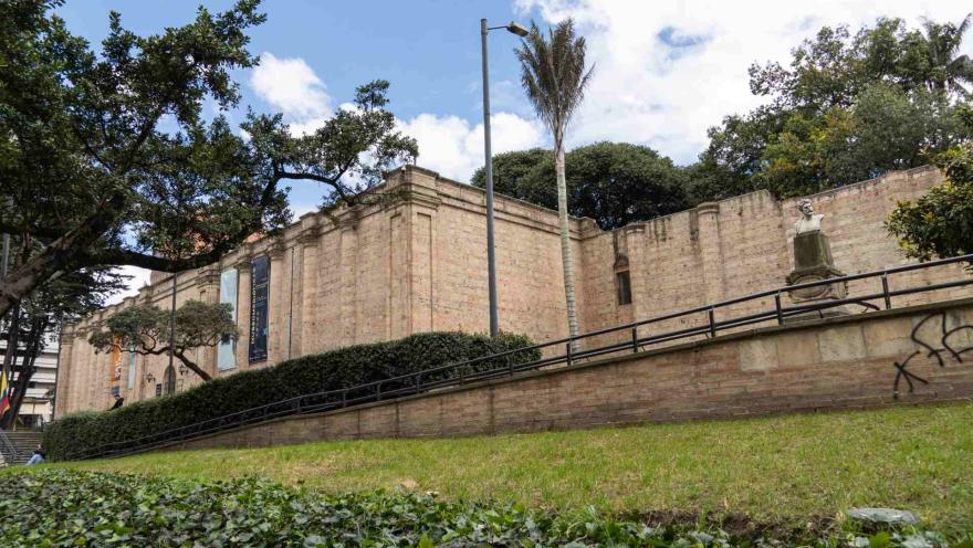 Museo Nacional de Colombia plano general de fachada visto desde abajo en la esquina izquierda, rodeado de vegetración, árboles y una alta palmera. Sobre el edificio de piedra hay un cielo azul con nubes. 