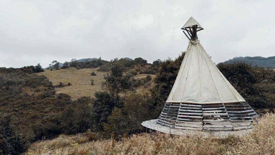 Páramo El Verjón, plano general vegetación en dos montañas con árboles secos y una estructura piramidal de madera como maloka blanca. El cielo es gris cubierto por nubes.