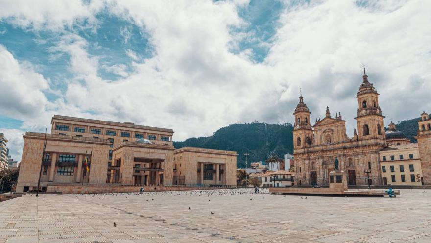 Plaza de Bolívar de día vista de esquina suroccidente al norte con palomas pequeñas a lo lejos, de frente el Palacio de Justicia, de lado la Catedral Primada, al fondo el cerro de Monserrate y el celo azul muy nubado.