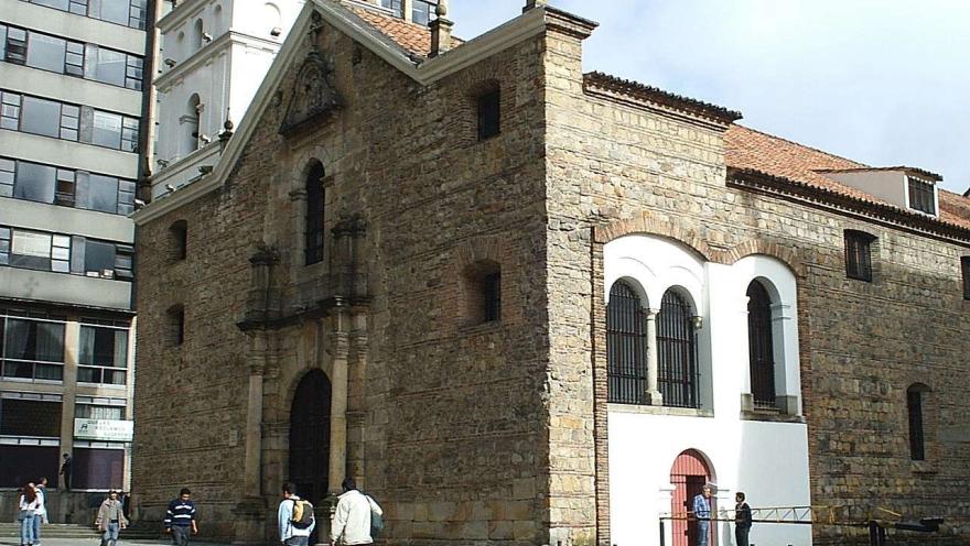  Templo de San Agustín colonial vista fachada y lateral izquierda, se observan gran puerta central, puerta lateral y ventanales. Al fono hay un edificio moderno. Algunas personas transitan por el frente.