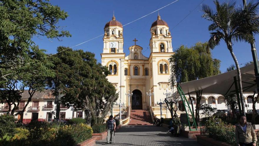  Basílica Menor de San Jacinto fachada de frente vista desde la paza rodeada de árboles y una palma, con un hombre con bastón y una mujer que caminan alejándose de ella. El cielo es muy azul.