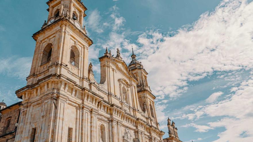 Catedral Primada de Colombia en primer plano contrapicado de la fachada  donde resaltan campanarios y torre del reloj. Se ve el cielo azul con nubes blancas tupidas.