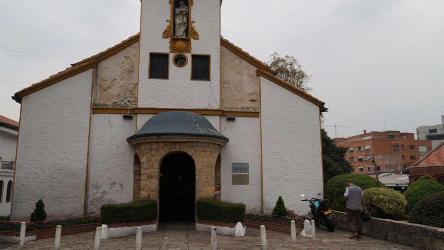 Iglesia Santa Mónica plano general fachada con un hombre adulto y una moto estacionada a la izquierda. Al frente el piso es en piedra y la puerta de la entrada está cerrada. al fondo a la izquierda aparece un edificio moderno.