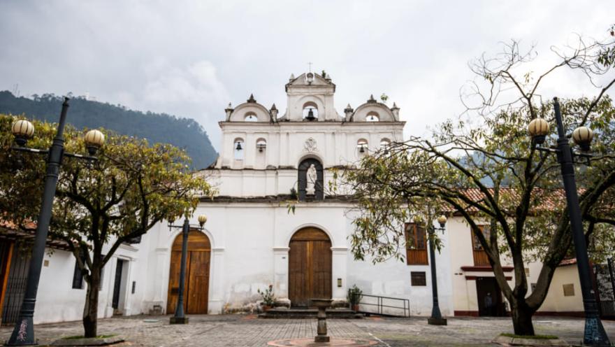 Parroquia de Nuestra Señora de Las Aguas fachada frente completa blancacon dos puertas gigantes en madera, dos ventanas, campanario con palomas y de fondo los cerros orientales. Ramas de árboles a los costados.