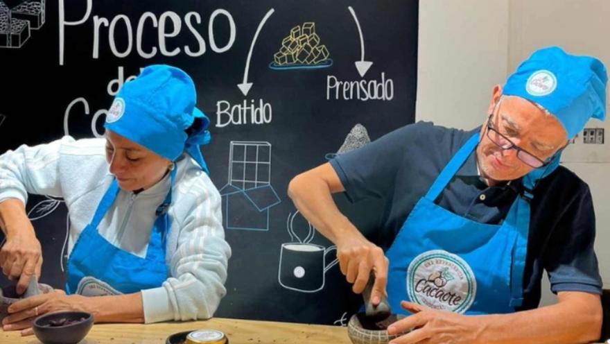 Un hombre y una mujer adultos con delantales y gorros azules muelen cacao en sendos morteros. Detás hay un aviso que señala el recorrido de la transformación del chocolate
