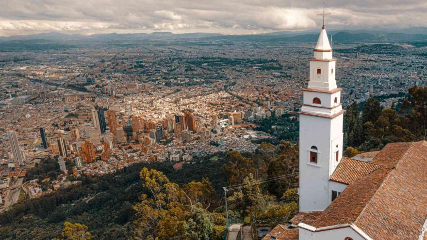 Monserrate Hill, close-up of the church tower and the mountain in a drone shot, with the vast city and its buildings stretching to the western mountain range in the background. The sky is overcast with clouds.