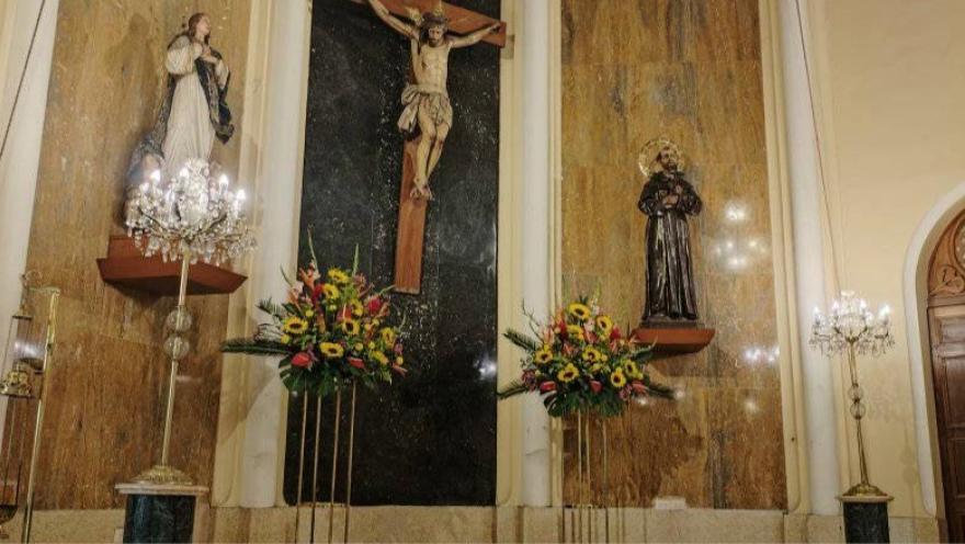  Iglesia La Porciúncula interior en el atrio con una imagen de Jesús crucificado con la Virgen a un lado y San Pedro al otro. Debajo de las figuras que reposan en la pared hay dos adornos florales y dos lámparas multi bombillos.