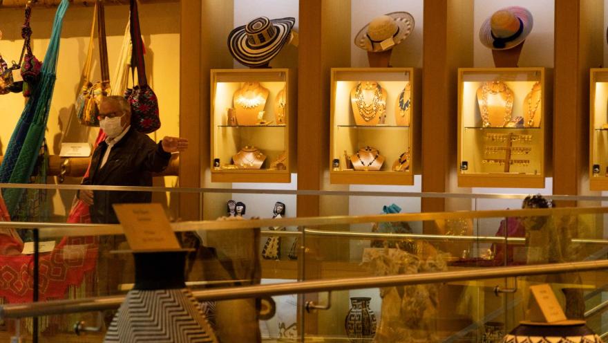 Museo del Oro en el salón de ventas con vitrinas en madera con objetos artesanales como sombreros, jarrones, cadenas, hamacas y bolsos. Un hombre adulto mayor con tabacoca señala con su mano izquierda.