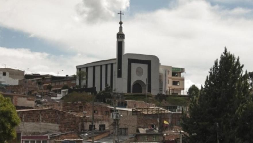 Santuario Nuestra Señora de la Peña vista de lejos en una cima detras de casas de barrio popular con cielo azul y nubes blancas. Destaca su fachada blanca con visos negros, su entrada en forma de óvalo y la torre con una punta alta que cierra con una cruz. 