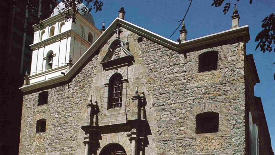 Templo de San Agustín vista de frente con fachada colonial en piedra gris, puerta en madera, ventanas con rejas y torre del campanario de color amarillo que termina en cúpula. decoran uns ramas de árbol y el cielo azul.