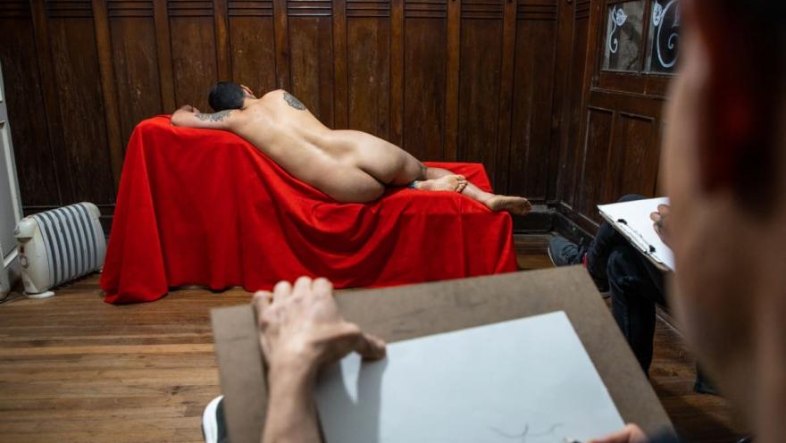 Chucuy Club clase de dibujo de espalda y torso desnudo de hombre que yace sobre un sofá cubierto con una tela roja. Hay un pintor mirando al hombre desnudo y haciendo trazos en papel.