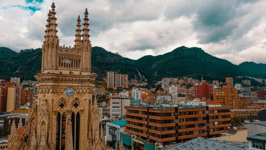 Iglesia Lourdes en primer plano la parte alta de la iglesia gótica con su torre del reloj y campanario, al fondo edificaciones incluso en los cerros orientales y un cielo nublado.