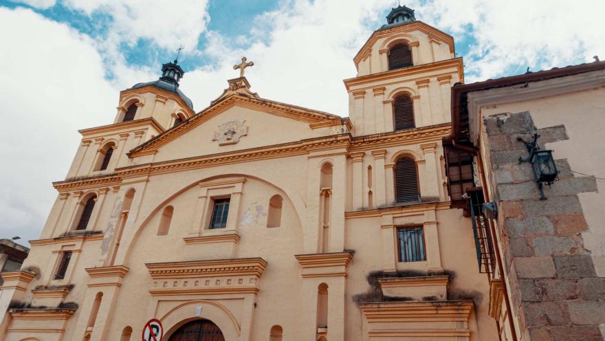  Iglesia de la Candelaria colonial de frente a su fachada, se ve desde la puerta de madera  con forma de óvalo hasta el techo coronado con una cruz y las torres izquierda y derecha con sendas cúpulas negras y cruces metálicas encima. 