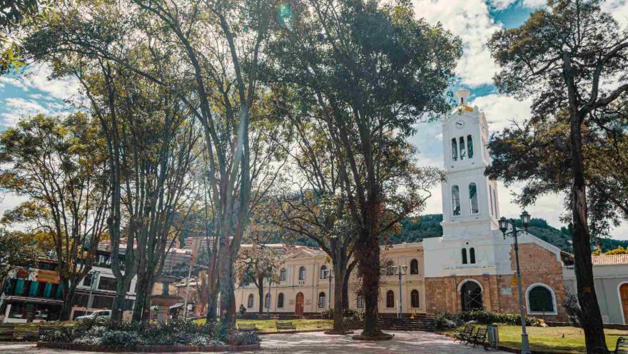 Plaza de Usaquén, plano donde se observa la iglesia, árboles altos y poco frondosos y de fondo los cerros orientales. El lugar está vacío.