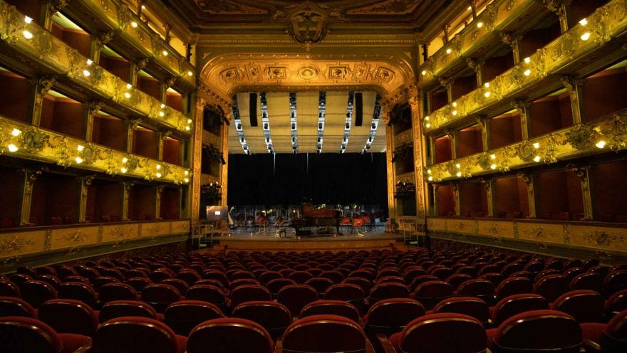 Teatro Colón interior, view from the last row of seats toward the stage, where there is a brown grand piano. The balcony lights are turned on.