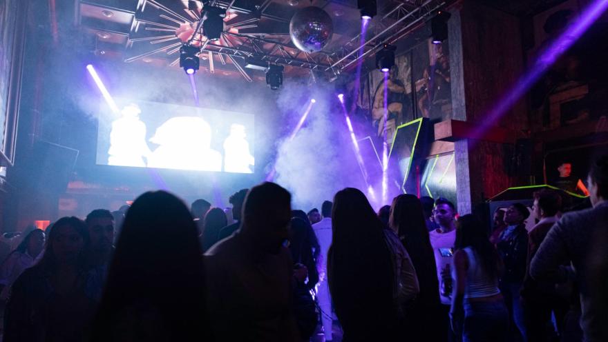 Theatron interior en sala de eventos oscura con iluminación de refletores, una pantalla gigante y sombras de varias personas que bailan y conversan