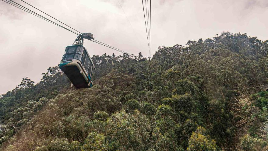 Cable car to Monserrate Hill, seen from below, hanging from cables above the mountain and its trees. The sky is cloudy.