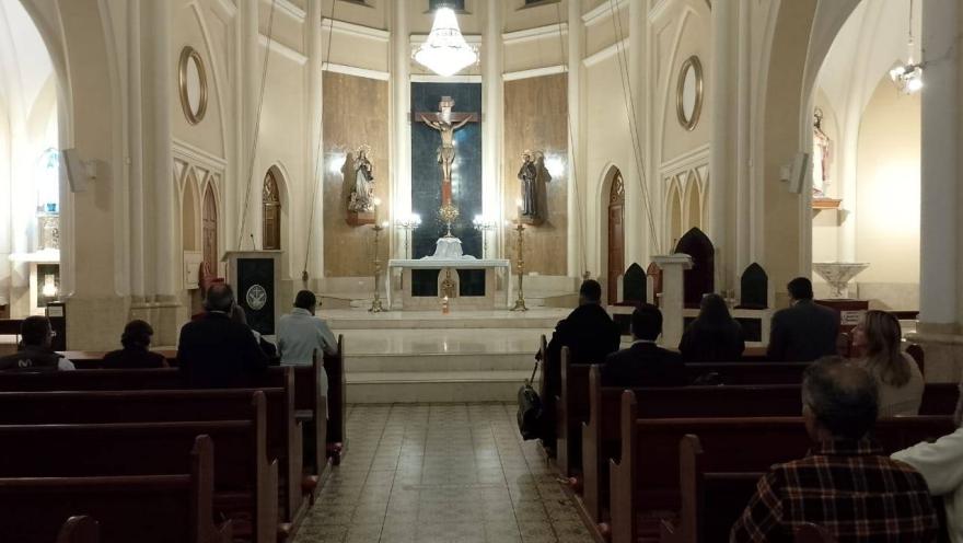  Iglesia La Porciúncula al interior personas orando sentadas y arrodilladas frente al altar. Destacan las paredes con círculos y óvalos y una gran lámapara que cuelga justo encima del Cristo.