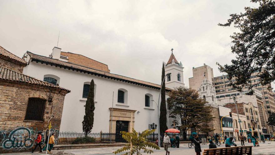  Iglesia de La Veracruz colonial vista lateral con dos pinos y árboles, y al frente la carrera séptima con algunos peatones caminando y otros sentados en sillas de madera. Hay algunas palomas y un perro blanco acostado en la vía.