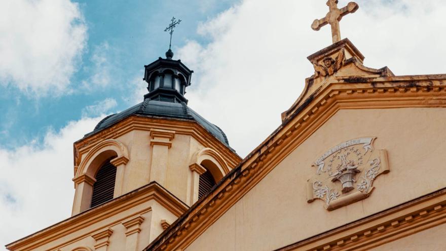  Iglesia de la Candelaria con la parte alta de su fachada en primer plano, se resalta la punta del frente coronada por una cruz y una torre con su cúpula negra y una cruz metálica encima. 