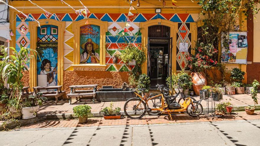  La Candelaria, fachada local de un piso con grafiti colorido en amarillo, azul y rojo con dibujo de campesinos. La puerta está abierta y al frente hay unas bicicletas estacionadas. Hay varias materas y plantas alrededor de la casa.