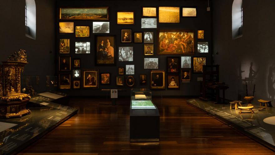 Museo Nacional de Colombia al interior sala de exhibiciones con una pared al fondo repleta de fotografías y cuadros. Acompañan la escena vitrinas que soportan artículos como sillas con valor histórico. El salón iluminado con luz artificial recibe también la natural desde dos pequeñas ventanas en la parte alta de los muros al lado izquiero y derecho.