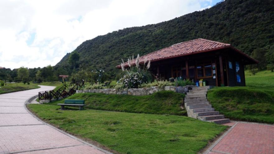 Parque Entrenubes con casa campestre moderna en madera y techos de teja de barro en la mitad de un camino de ladrillo, está rodeada por cesped, una silla en metal y concreto, plantas pequeñas y una escalera en piedra. Al fondo se ve la montaña y un cielo azul cubierto por nubes.