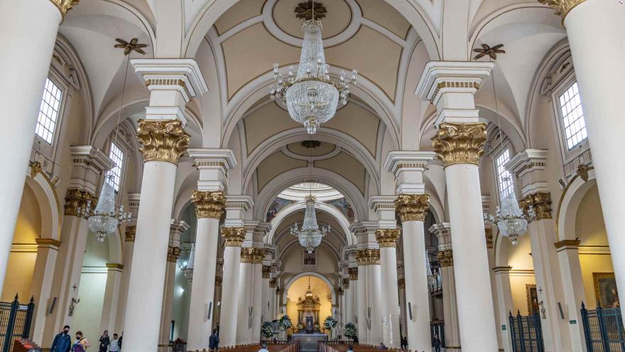 Catedral Primada de Colombia, interior vista desde la puerta hacia el altar de fondo con muchas filas de sillas con pocas personas sentadas. Destacan 7 grandes columnas blancas a cada lado, los ventanales y 2 gigantes lámparas que cuelgan del techo. 