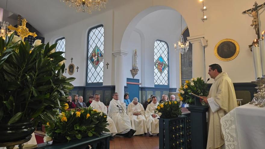  Iglesia Santa Mónica al interior primer plano del sacerdote en el atrio que habla al microfono para un grupo de sacerdotes. Lo rodean cuatro arreglos florales, un Cristo a espaldas, una Virgen María al frente y dos lámparas que cuelgan del techo.