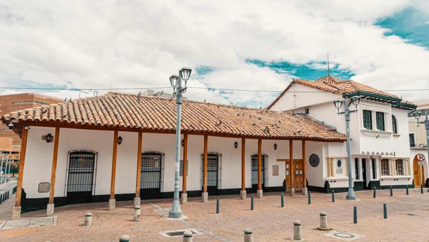 Plaza de Usaquén, priemr plano de casa esquinea antigua con fachada blanca, techo en teja y soportado por parales de madera café. En la calle enladrillada al frente hay bolardos y dos faroles gigantes. El cielo azul está casi cubierto por nubes.