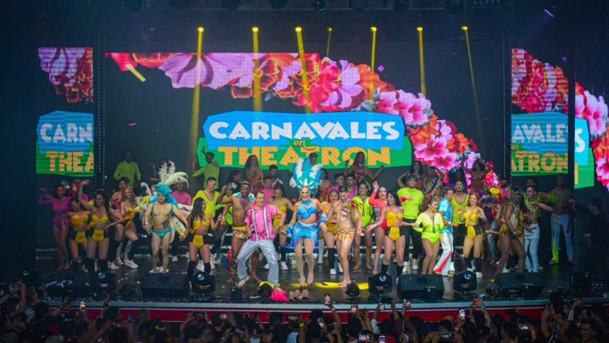 Theatron vista clara de escenario con decenas de artistas disfrazados con trajes de colores y un título que dice Carnavales en Theatron. Al frente de la tarima se alcanza a ver a un grupo de personas que levantan la mano.