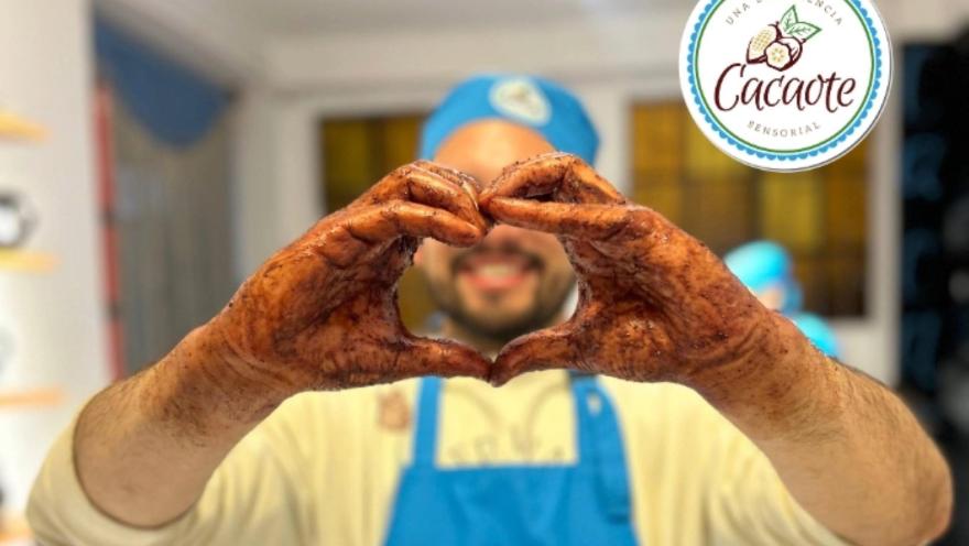 Un hombre joven con delantal y gorro azul muestra sus manos untadas de cacao y haciendo una forma de corazón.