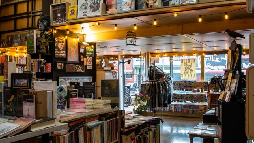 La Valija de Fuego interior del local con estanterías repletas de libros, incluso en las paredes, y un exhibidor de ropa, parches y objetos decorativos. Hacia afuera se ve la calle con luz de día.