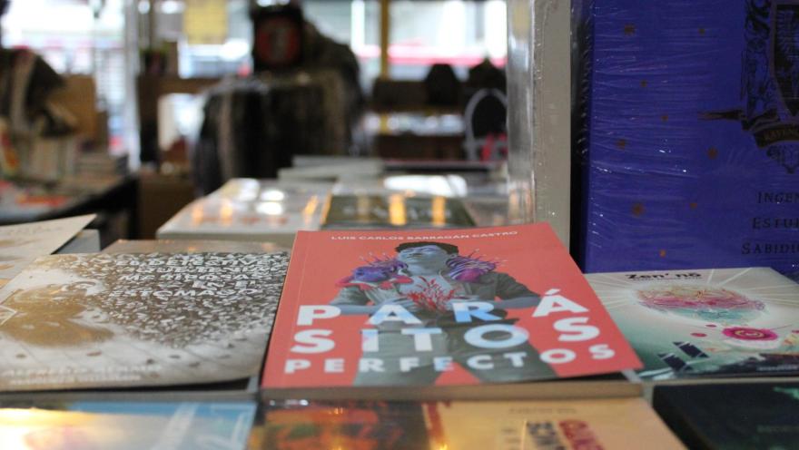  Librería La Valija de Fuego primer plano sobre libros en una mesa en el que se destaca uno rojo cuyo título es Parásitos perfectos, al fono borrosa una ventana con luz de la calle.