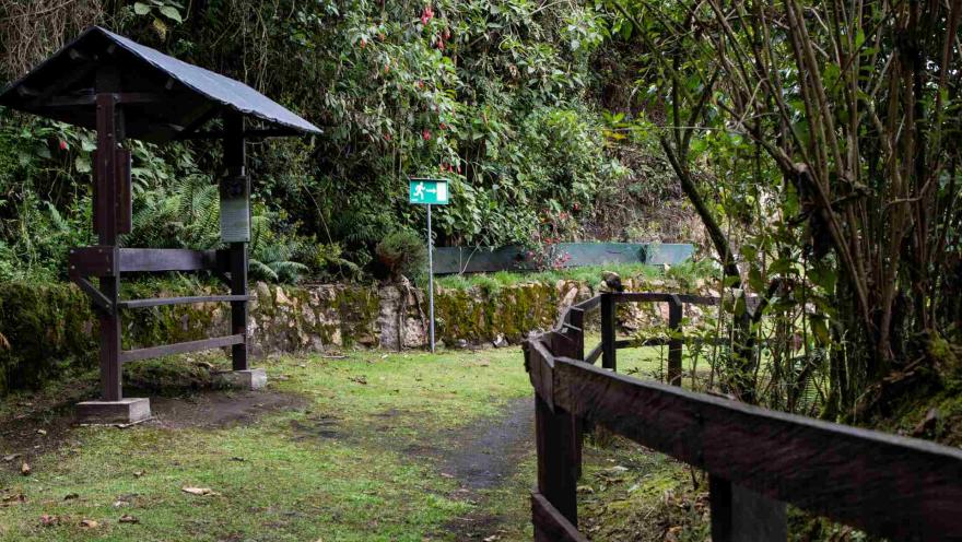  Sendero El Paramuno en curva de camino de tierra con hierba demarcado con una cerca de madera y unas rocas grandes cubiertas por musgo. Hay dos señalizaciones, una en madera, cubierta con teja para cubrirse de la lluvia y otra metálica de color verde que orienta la direccion a seguir.