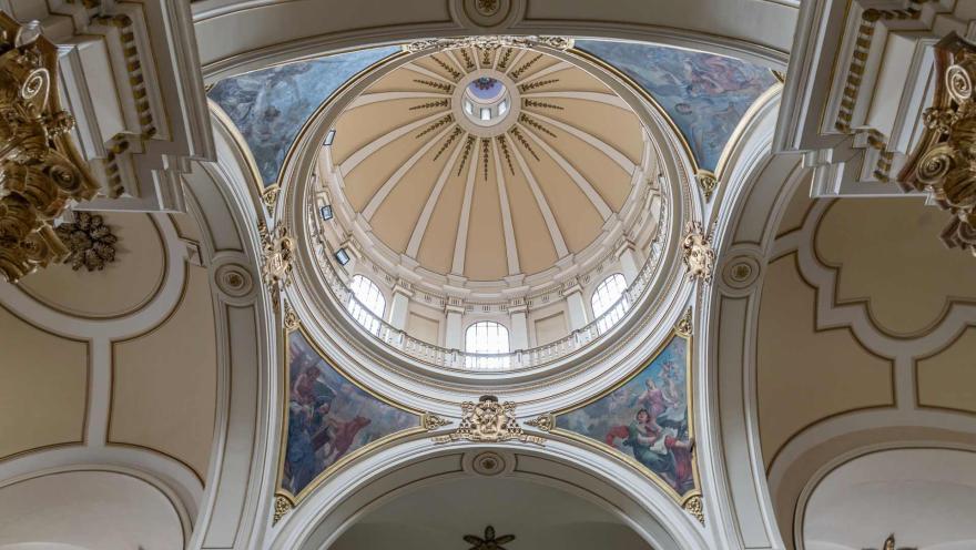 Catedral Primada de Colombia, cúpula del atrio vista desde abajo en la que destaca la fina mampostería y los murales entre azules y rojos con figuras de personas. La cúpula destaca el decorado en forma de estrella y 3 ventanales con luz blanca.