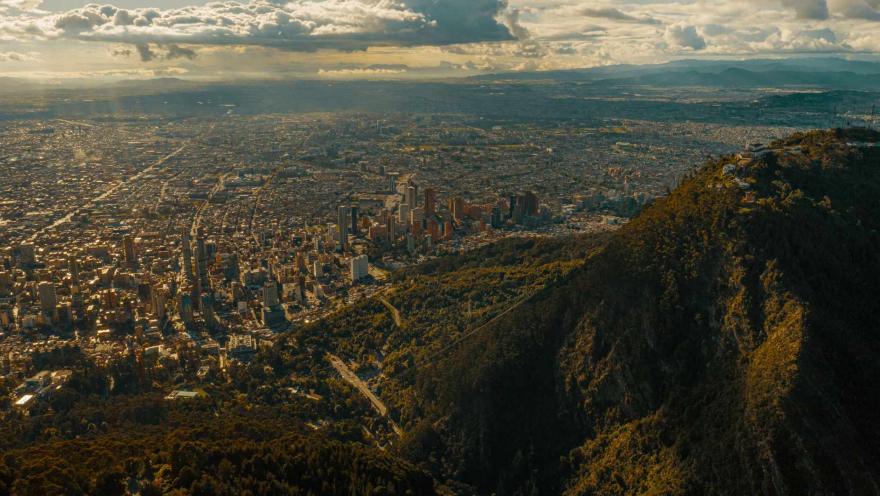 Cerros Orientales con la ciudad vista en picada desde la cima de una montaña. El cielo es azul con nubes y haces de luz solar.
