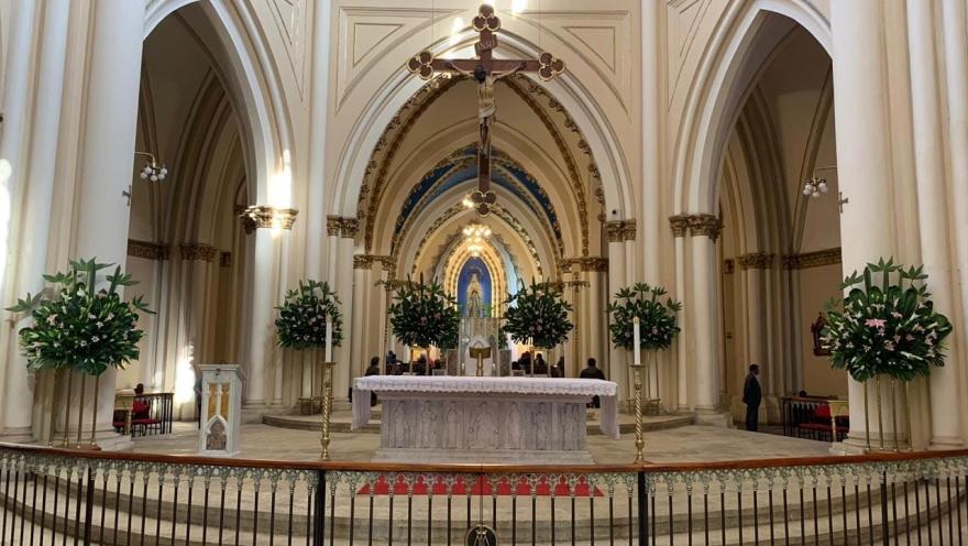 Iglesia Lourdes al interior plano general del altar con sus paredes ovaladas y un Cristo en el centro, al fondo la Virgen con personas que le oran. El altar está decorado con arreglos florales y dos velones. 